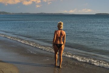 A slender blonde girl in a bikini is alone on a sandy beach in Athens, Greece on a summer day, photo at sunset from the back.