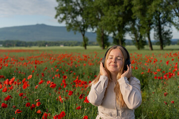 Girl with headphones listening to music on a blurred green background of poppy field and mountains. Woman feels happy in nature. European woman dancing and listening to music with headphones. 