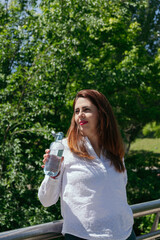 Portrait of a mature woman with long hair and red lips in nature drinking a bottle of water after a...