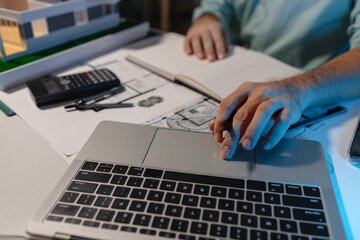 an engineer is doing a big project in the office, an architect uses electronic device to make the work he is doing easier, blueprint and model are on the desk, employee is working with concentration