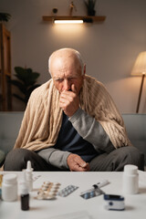 Senior man sitting sick on sofa at home, coughs, covers his mouth with his hand. Suffers from...