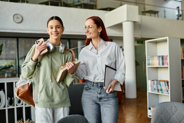 A redhead woman tutors a teenage girl in a library, both holding books, engrossed in after-school...