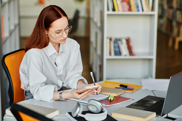 A redhead female tutor sitting at a desk, teaching online with a laptop and cell phone.