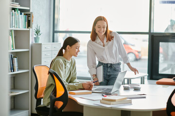A redhead tutor guides a teenage student through after-school lessons at a desk in a modern office,...