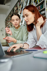 A tutor with red hair guides a teenage student in a library, both engrossed in the laptop screen for after-school lessons.