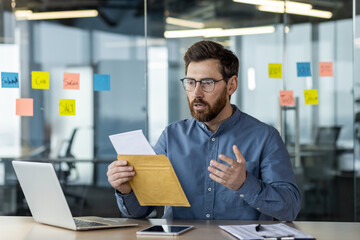 Worried young man sitting in office at desk with laptop and reading received letter and documents in shock