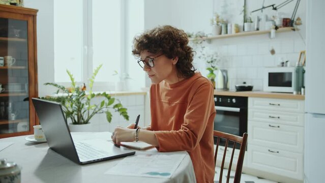 Young woman typing on laptop, taking notes and analyzing financial documents while working remotely from home