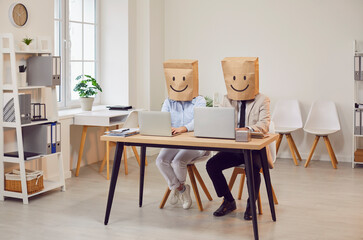 Business coworkers working together with laptops in office, wearing a paper bag on head depicting positive emotions and smiles. Maintaining positivity and a cheerful attitude in the workplace.