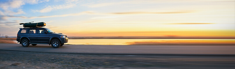 SUV with a kayak on the roof rack driving along a scenic lakeside road at sunset, reflecting an...