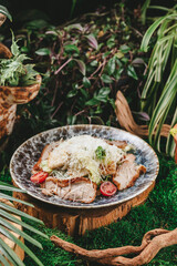 Close-up of a delicious Caesar salad with grilled chicken, Parmesan cheese, and cherry tomatoes, served on a blue marble platter in a lush green background setting.