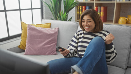 Fototapeta premium A smiling mature hispanic woman enjoying leisure time in a cozy living room with a remote control.
