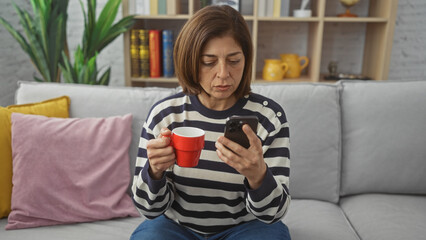 Obraz premium A middle-aged hispanic woman in a striped sweater sips coffee while checking her smartphone, seated on a gray couch indoors.