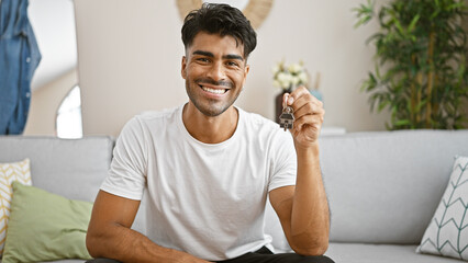 A smiling young hispanic man holding house keys indoors, conveying a sense of homeownership or...