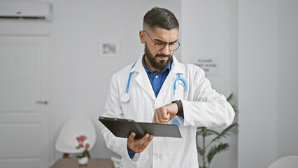 A handsome man wearing a labcoat checks the time while holding a tablet in a modern clinic's...