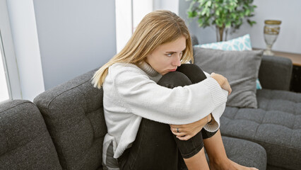 A contemplative blonde woman sitting indoors on a sofa in a cozy room, evoking a sense of...