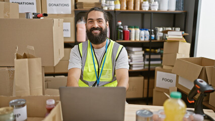 Handsome bearded man smiling at workplace with laptop, donation boxes, and safety vest in a...
