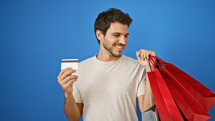 A smiling young hispanic man holding a credit card and shopping bags against a blue background.