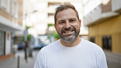 Fototapeta premium Handsome mature hispanic man with grey hair smiling confidently on a sunny urban street.