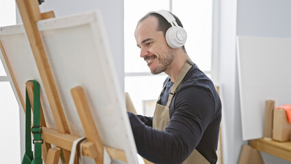 A bald bearded man wearing headphones paints on an easel in a bright art studio.