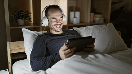 Smiling bearded man using tablet with headphones in bed at night, reflecting modern leisure or relaxation at home.