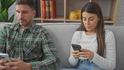 A man and woman use smartphones separately on a sofa in a cozy living room, implying disconnect...