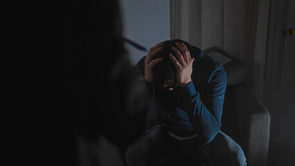 Obraz premium A distressed man sits indoors with his head in his hands, suggesting an interrogation or a dramatic scene.