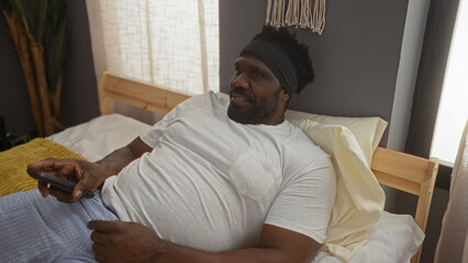 Fototapeta premium A young, handsome african american man with a beard relaxes in bed at home, holding a remote, in a cozy and well-lit bedroom.