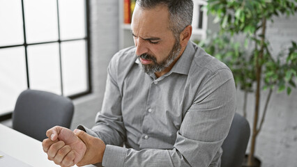 A mature man with grey hair and beard feels hand pain in an office setting.