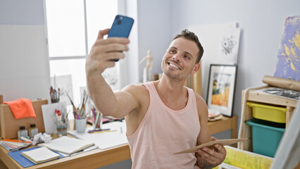 A smiling hispanic man takes a selfie in a bright art studio holding a paintbrush.