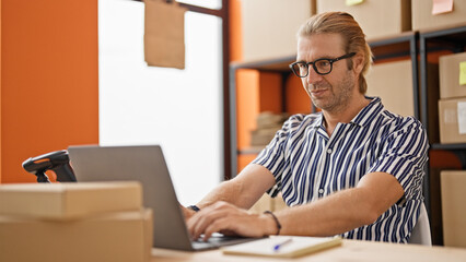 Obraz premium A blond businessman with glasses working on a laptop in a warehouse office.