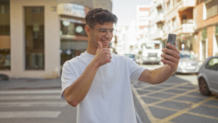 Young hispanic man taking a selfie on an urban street while giving a thumbs-up, wearing glasses and a white t-shirt.