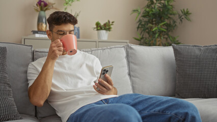 A young hispanic man relaxing on the sofa in his living room, holding a smartphone and sipping...