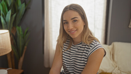 A beautiful blonde woman sits on a bed in a cozy bedroom, wearing a striped shirt and smiling warmly, with a lamp and plant in the background adding to the comfortable home setting.
