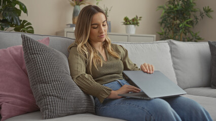 Obraz premium Attractive young woman with blonde hair using a laptop while sitting on a cozy sofa in a modern living room with indoor plants in the background.