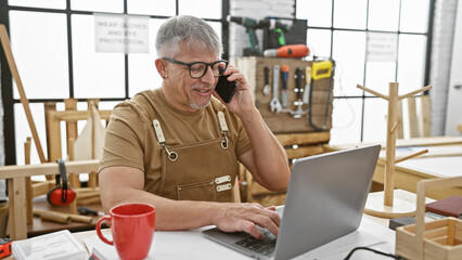 Grey-haired man in apron using laptop and phone in a sunny carpentry workshop