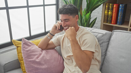 A cheerful young hispanic man chatting on a smartphone while sitting on a sofa indoors with colorful cushions and a bookshelf