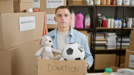 Young hispanic man standing in a warehouse surrounded by donation boxes containing toys and...