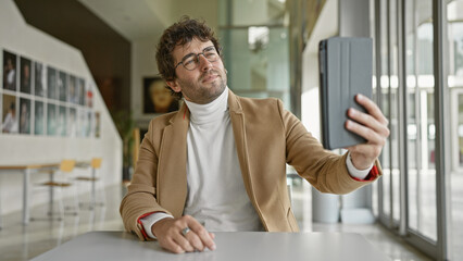 Fototapeta premium Handsome hispanic man with beard taking a selfie indoors at a modern office, wearing glasses and a casual turtleneck and blazer.