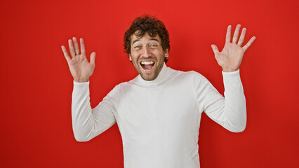 Naklejka premium A joyful hispanic man in a white sweater posing with raised hands against a vibrant red wall.