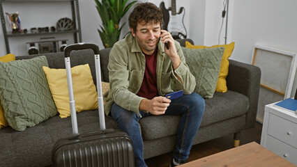 Obraz premium Handsome hispanic man with beard talking on phone and holding credit card, sitting on couch at home with suitcase.
