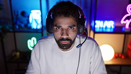 Hispanic man wearing headset in a dark gaming room at night with colorful neon lights.
