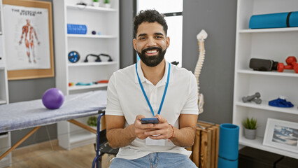Handsome hispanic man with beard smiling in a physical therapy clinic room holding a smartphone,...
