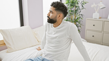 Obraz premium A contemplative young hispanic man with a beard sits thoughtfully in a bright bedroom interior.