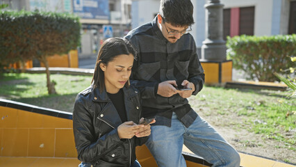 A man and a woman focused on smartphones while sitting together on a bench in a sunny urban setting