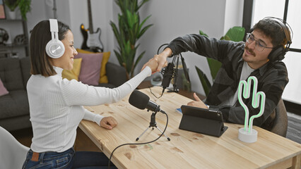 A woman and a man in a podcast studio, shaking hands over a wooden table with microphones and...