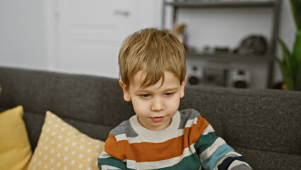 Fototapeta premium Caucasian toddler boy sitting thoughtfully on a grey couch inside a cozy living room