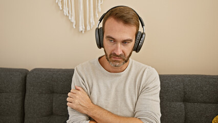 A pensive middle-aged man with a beard wears headphones in a home interior, evoking telecommuting...