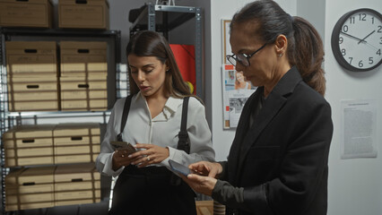 Two women in an office setting, one examining a smartphone and the other looking at papers,...