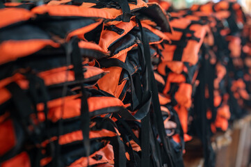 Many orange and black life jackets stacked together. It is placed in a storage area. They are used for water-related activities or for emergency preparedness.