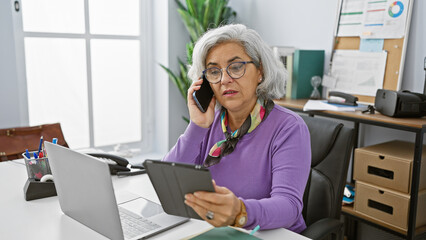 A mature woman multitasks in a modern office, using a smartphone and tablet with a laptop and...